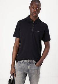 Melna polo krekls ar rāvējslēdzēja apkakli un logotipa akcentu uz krūšu, apvienots ar gaiši pelēkiem džinsiem, turēts ar melnu somu.