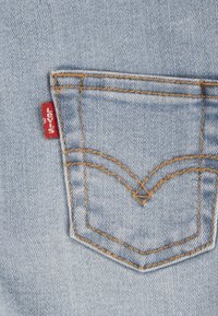 Ljusblå denimficka med gul sömnad, som har en röd etikett med vit text. Klassisk design med en rundad topp och spetsig botten.