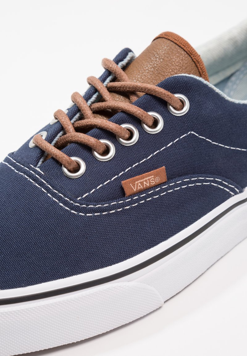Zapatillas VANS con parte superior de lona azul marino, acento de cuero marrón, suela de goma blanca y cordones marrones a través de ojales plateados. Detalle de costura blanca.