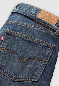 Poche de jean en denim bleu avec coutures orange, étiquette en tissu rouge Levi's, et étiquette de marque en cuir marron Levi Strauss & Co. au-dessus de la ceinture.