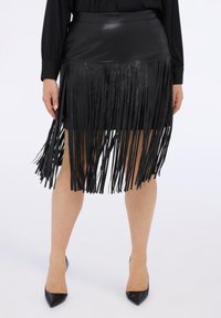 Czarna skórzana spódnica z frędzlami o dopasowanej talii i teksturowanym wykończeniu; asymetryczne frędzle sięgają poniżej linii dołu, w zestawieniu z czarnymi, spiczastymi obcasami.