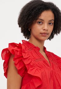 Rød bluse med volanger på ermene, teksturert stoff og blonder detaljer ved halsen, med en avslappet passform.