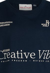 Navyblauwe katoenen t-shirt met witte geborduurde tekst: "International Dreamers," "Creative Studio," en "Follow Freedom - Retour Jeans."
