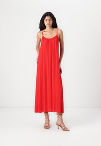 Robe maxi rouge avec des bretelles fines, en matériau léger, coupe décontractée, et encolure élastique. Associée à des sandales à talons hauts nude à brides.