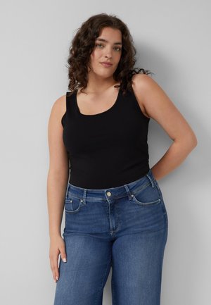 Frau mit lockigem dunklem Haar, die ein schwarzes ärmelloses Oberteil und blaue Jeans trägt, vor einem schlichten hellgrauen Hintergrund stehend.