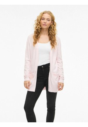 Donna con capelli biondi ricci che indossa un cardigan rosa chiaro, una canottiera bianca e jeans neri, in piedi davanti a uno sfondo bianco semplice.