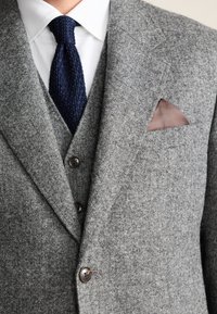 Blazer en laine grise avec une finition texturée, doté d'une fermeture à deux boutons, d'une cravate en tricot bleu marine et d'un mouchoir de poche marron à motifs.