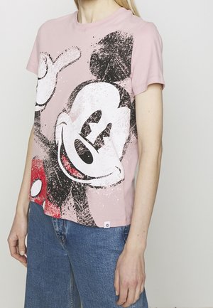Osoba nosząca różowy T-shirt z dużą, postarzaną grafiką Myszki Miki i niebieskie dżinsy, stojąca na tle prostego jasnego tła.