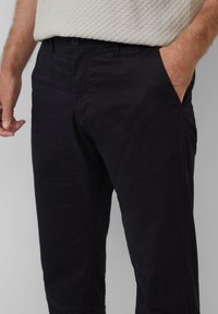 Pantalon en coton noir avec une texture lisse, doté d'une fermeture à bouton et de poches latérales, partiellement visible sur un fond gris clair.