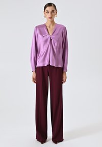 Donna che indossa grandi orecchini d'oro, una blusa lucida color lavanda con un nodo frontale e pantaloni larghi borgogna, in piedi contro uno sfondo semplice.