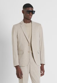 Completo in lino beige chiaro con giacca strutturata e pantaloni coordinati. Abbinato a una maglietta beige e occhiali da sole neri oversize.