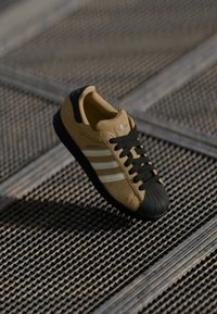 Brown suede sneaker with black rubber toe cap, black laces, silver three-stripe design, and logo on the tongue. Textured surface beneath.