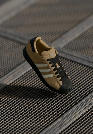 Brune semskede sneakers med sort gummitåkappe, sorte snørebånd, sølvtre-stripes design og logo på tungen. Tekstureret overflade nedenfor.