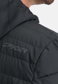 Schwarze Pufferjacke mit Kapuze, ausgestattet mit einem gesteppten Design und einem in einem dunkleren Farbton sichtbaren "SPYDER"-Logo im Bereich der linken Schulter.
