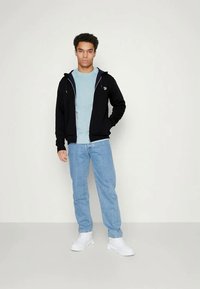 Giovane uomo in piedi con una mano nella tasca di una felpa nera con zip, indossa una maglietta azzurro chiaro, jeans blu e sneakers bianche, su sfondo neutro.