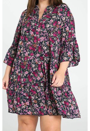Vrouw draagt een donker gebloemde jurk met roze en witte bloemen, driekwartmouwen met ruches en een losse, gelaagde rok.