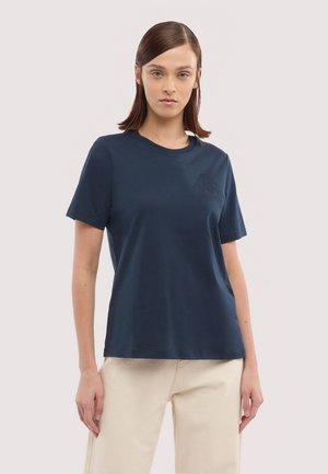 Kobieta z prostymi brązowymi włosami, ubrana w granatowy t-shirt z krótkim rękawem i jasnobeżowe spodnie, stojąca na neutralnym tle.