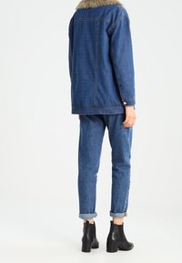 Riflová bunda s umělým kožíškem na límci, modré džínové kalhoty vyhrnuté na kotnících a černé kotníkové boty. Oblečení má volný střih a texturovanou tkaninu.