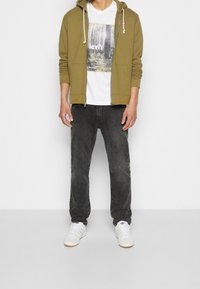 Sudadera con cremallera en verde oliva, camiseta gráfica blanca con diseño de árbol, jeans oscuros grises, zapatillas blancas con acentos en negro y marrón.