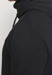 Felpa nera con colletto alto, realizzata in tessuto morbido. Presenta una tasca frontale, corde di regolazione e una vestibilità comoda. Texture liscia e design minimalista.