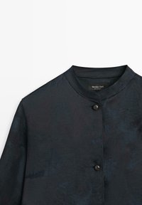 Blouse à manches longues bleu foncé, avec un col rond, une finition subtilement texturée et une fermeture à bouton unique à l'avant. Étiquette de la marque visible.