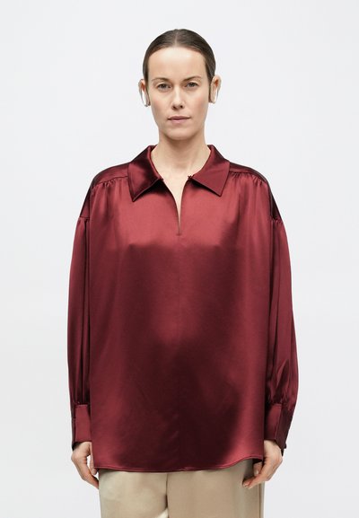 Kvinde med glat hår iført beige bukser og en løstsiddende, langærmet, bordeauxfarvet satinskjorte mod en ensfarvet baggrund.