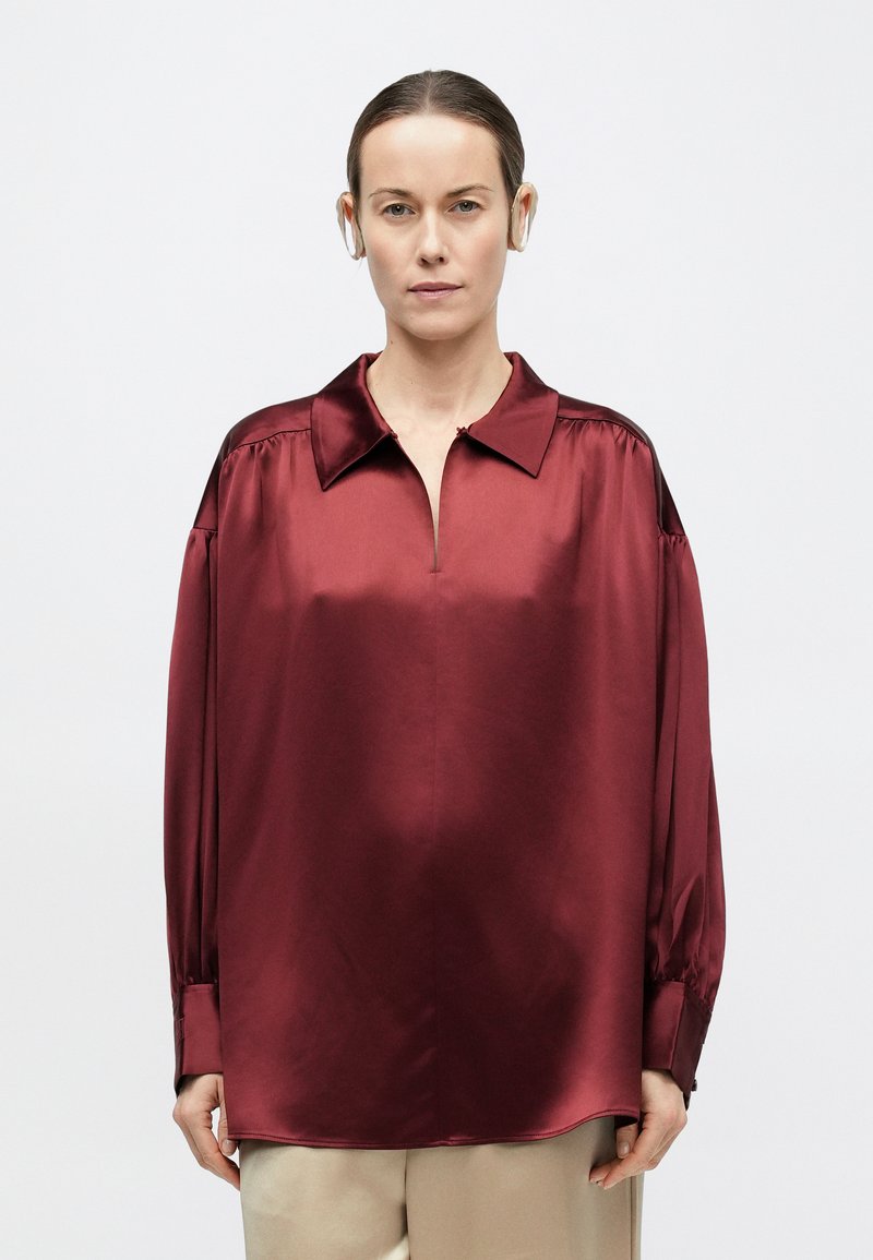 Woman with straight hair wearing beige pants and a loose-fitting, long-sleeve, burgundy satin shirt against a plain background.