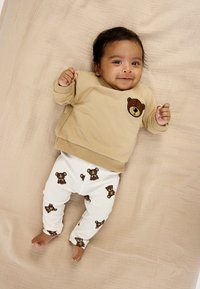 Beige sweatshirt med bjørneemblem, kombineret med hvide ribbukser med brune bjørneprint. Blødt stof på en tekstureret beige baggrund.