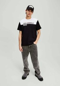 Mustvalge lühikeste varrukate T-särk, millel on rinnal trükitud "KARL LAGERFELD JEANS", koos kulunud hallide pükste ja tumedate kingadega.