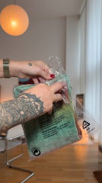 Barvna, teksturirana tkanina ovita v prozorno plastično embalažo. Opazite vidne simbole za recikliranje in navodila za nego na hrbtni strani.