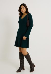 Robe verte à col en V côtelé avec des manches longues et un ourlet évasé, associée à des bottes en cuir marron jusqu'au genou et un petit sac sombre.