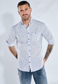 Hellgraues, gestreiftes Button-Up-Hemd mit hochgekrempelten Ärmeln, schwarzen Knöpfen und einer Brusttasche. Kombiniert mit blauen Jeans mit glatter Textur.
