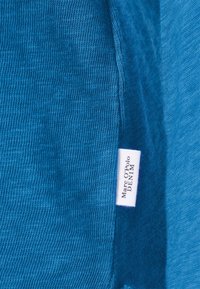 Blauwe textuurstof met een ribbeloppervlak, waarop een wit label met "Marc O'Polo DENIM" aan de zijkant is te zien.