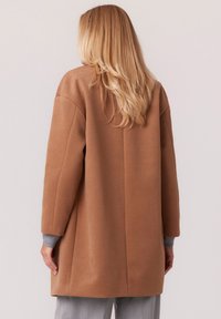 Brązowy, teksturowany płaszcz o luźnym kroju, ze zredukowanymi ramionami i długimi rękawami. Szare mankiety widoczne przy nadgarstkach. Prosty, czysty design.