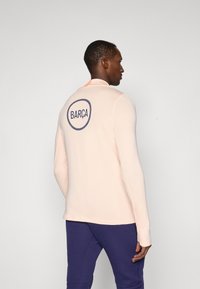 Long-sleeve, light peach athletic top with a rounded navy "BARÇA" logo on the back. Features a zip collar and fitted design.