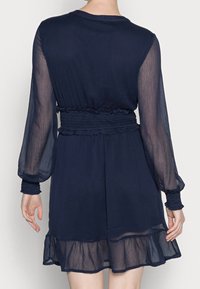 Mörkblå klänning med långa transparenta ärmar, smockad midja och volangkant. Lätt, luftig tyg med en texturerad yta.