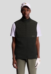 Gilet sans manches noir avec devant matelassé, col montant et un logo subtil. Porté sur une chemise blanche, complété par un pantalon gris et une casquette.