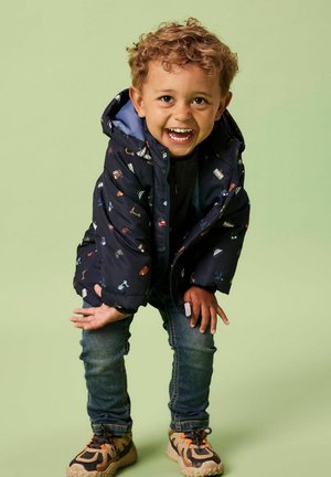 Giubbotto piumino blu navy con motivi colorati di cartoni animati, con cappuccio, indossato sopra una maglietta scura, abbinato a jeans blu e sneakers marroni.