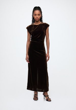 Femme aux cheveux tressés portant une robe maxi en velours marron foncé sans manches et des sandales noires à talons hauts, debout devant un fond uni.