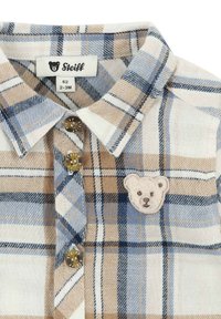 Camisa a cuadros en beige, azul y blanco. Presenta un parche de oso en el pecho y botones dorados. Tejido suave con cuello tipo polo.