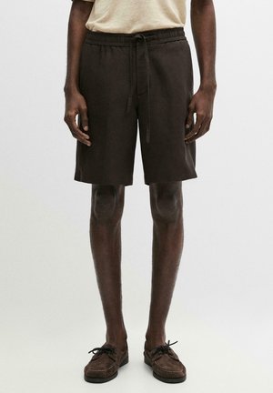 Uomo con pantaloncini scuri con coulisse, maglietta beige e mocassini marrone scuro in piedi davanti a uno sfondo chiaro e uniforme.