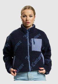 Ženska z modrim flis jopičem in svetlom modrim zadrgo ter žepom na prsih, ki stoji ob lahkem enobarvnem ozadju.