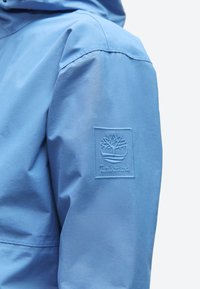 Μανίκι ανοιχτό μπλε μπουφάν με ραμμένη την ετικέτα Timberland, που απεικονίζει ένα δέντρο μέσα σε τετράγωνο στο πάνω μέρος του μπράτσου.