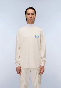 Ilgas rankovėmis smėlio spalvos marškinėliai iš minkštos medžiagos, su mažu daugiabareviu piešiniu kairėje krūtinės pusėje, derinami su šviesios spalvos kelnėmis.