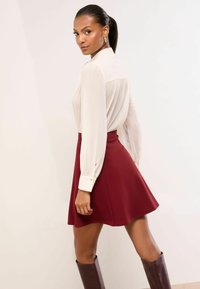 Naine, kellel on tagasitõmmatud juuksed, kannab kreemika värvi pika varrukaga pluusi, punast lehvivat seelikut ja pruune põlviksaapaid ning vaatab üle õla.