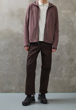 Haut à capuche zippé bordeaux avec une capuche, polo blanc en dessous, pantalon marron et chaussures de sport noires. Tissu texturé, coupe décontractée.