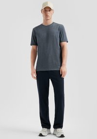 T-shirt noir gris à manches courtes, décolleté ras du cou, associé à un pantalon bleu marine et des baskets gris clair, portant une casquette beige. Design simple.