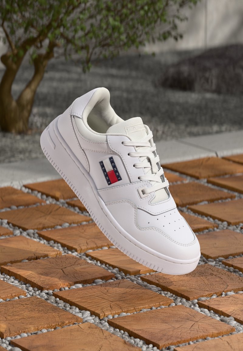 Baskets basses blanches avec logo Tommy Jeans sur le côté, flottant au-dessus d'une tuile en bois et d'une surface extérieure en galets avec un arrière-plan flou d'arbres.
