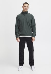 Green fleece pullover with a quarter-zip neck, worn over a white shirt, paired with black pants and white sneakers. Simple, textured design.