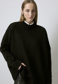 Žena s ravnom plavom kosom, koja nosi srebrne naušnice u obliku prstena, preveliki crni džemper s rupama i crne kožne hlače, stoji protiv jednostavne pozadine.